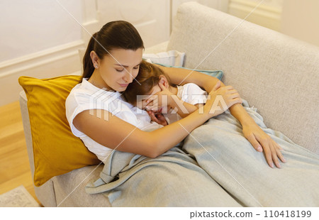 Mother embracing sleepy child while they relaxing on cozy sofa at home 110418199