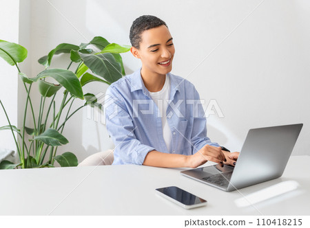 Positive millennial latin lady with phone with empty screen at table typing on computer 110418215