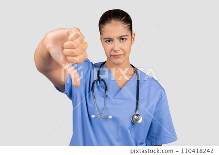 A European millennial doctor in blue scrubs shows disapproval with a thumbs-down A European millennial doctor in blue scrubs shows disapproval with a thumbs-down 110418242