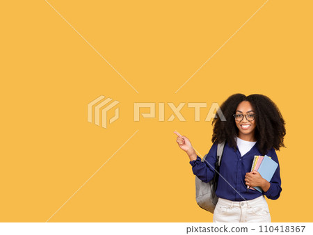 Happy black lady student in glasses, with backpack and copybooks, pointing aside at free space, on yellow background 110418367
