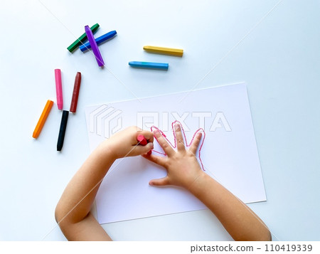 Childrens hands draw their hand with wax crayons on white paper, top view. Childrens hands draw their hand with wax crayons on white paper, top view. 110419339