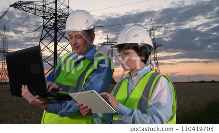 Engineers compare information on computers by power transmission lines at sunset Engineers compare information on computers by power transmission lines at sunset 110419703