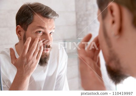 Portrait of man in white tshirt applying cream on his face at bathroom. Self care morning procedure. 110419997