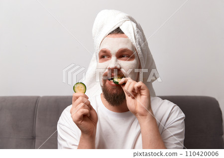 Portrait of funny happy man in white tshirt applying cucumber slices over clay mask on his face. Self care morning spa procedure Portrait of funny happy man in white tshirt applying cucumber slices over clay mask on his face. Self care morning spa procedure 110420006