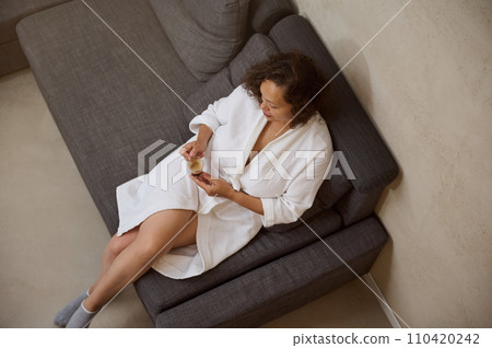 Top view curly haired brunette, young pretty woman in white bathrobe, sitting on couch at home and drinking coffee. 110420242