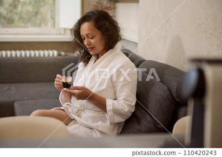 Happy curly haired brunette woman drinking cup of tea in an autumn morning. Dreaming lady in white bathrobe, sitting in living room with cup of hot aroma espresso coffee, enjoying happy weekend indoor Happy curly haired brunette woman drinking cup of tea in an autumn morning. Dreaming lady in white bathrobe, sitting in living room with cup of hot aroma espresso coffee, enjoying happy weekend indoor 110420243