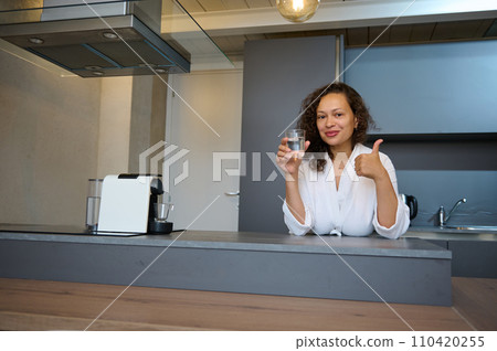 Beautiful curly woman smiles, thumbs up looking at camera, holds a glass of mineral water, standing at kitchen counter 110420255