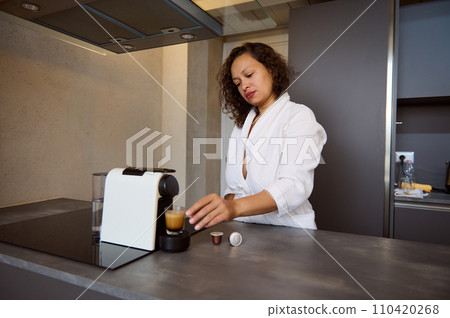 Portrait of a sleepy woman in white bathrobe, using capsule coffee machine, makes her morning espresso at her kitchen 110420268