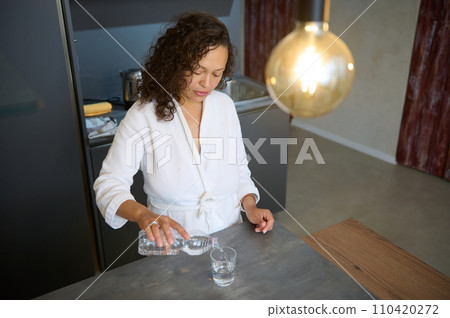 Young woman standing at kitchen counter in modern minimalist home interior, pouring some water into a drinking glass 110420272