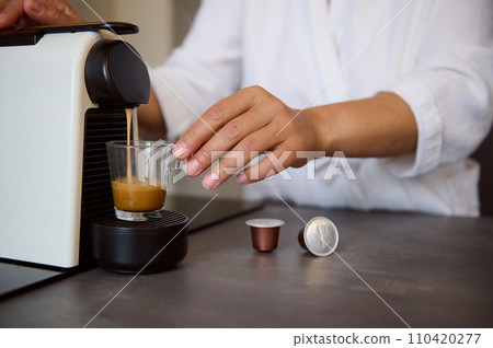 Close-up hands of housewife using a capsule coffee machine preparing freshly brewed espresso coffee in cozy home kitchen 110420277