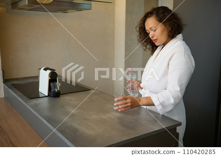 Authentic middle aged multiracial woman in white waffle bathrobe starts her day with a glass of rehydrating fresh water 110420285