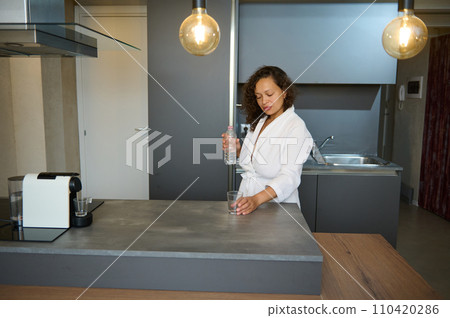 Beautiful African American sleepy woman in white robe holding a glass of refreshing water, standing at kitchen island Beautiful African American sleepy woman in white robe holding a glass of refreshing water, standing at kitchen island 110420286