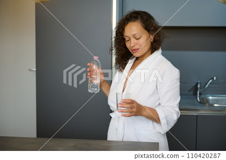 Multi ethnic pretty woman standing at kitchen counter, dressed in white bathrobe, starting her day with glass of water 110420287