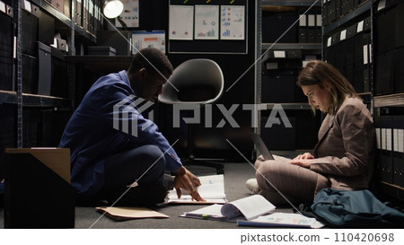 African american investigator assists with case files in evidence room as policewoman searches online for criminal records on laptop. Private detective thrilled to provide important information. 110420698
