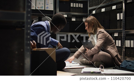 Black policeman and female law officer sitting on office floor with paperwork and laptop as crime investigation is being conducted. Multiracial investigators review and debate case evidence and hints. Black policeman and female law officer sitting on office floor with paperwork and laptop as crime investigation is being conducted. Multiracial investigators review and debate case evidence and hints. 110420700
