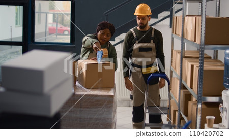 Manager wearing protective equipment in warehouse supervising african american trainee sealing cardboard box packages. Storage area personnel properly securing goods to avoid problems during shipping Manager wearing protective equipment in warehouse supervising african american trainee sealing cardboard box packages. Storage area personnel properly securing goods to avoid problems during shipping 110420702