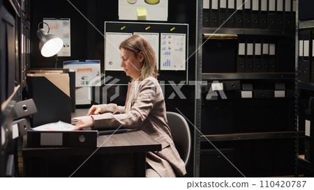 Caucasian detective inspector examines case files while seated at desk with laptop. Female private investigator carrying backpack enters police department office space and goes through crime records. Caucasian detective inspector examines case files while seated at desk with laptop. Female private investigator carrying backpack enters police department office space and goes through crime records. 110420787