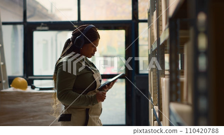 Happy warehouse head of logistics listening music while inspecting merchandise. Cheerful african american director wearing heaphones while walking through storage establishment rows Happy warehouse head of logistics listening music while inspecting merchandise. Cheerful african american director wearing heaphones while walking through storage establishment rows 110420788