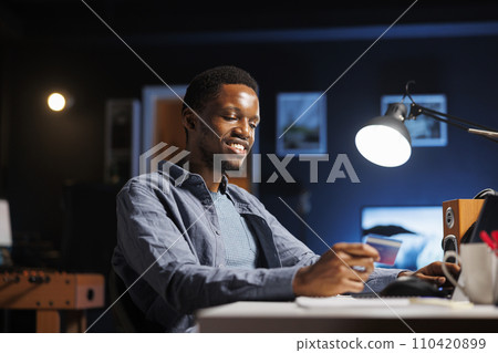 Pleased freelancer doing online shopping session on laptop, adding banking credentials to make transaction. Young happy man buying clothes during sale season, working from home on new project. 110420899