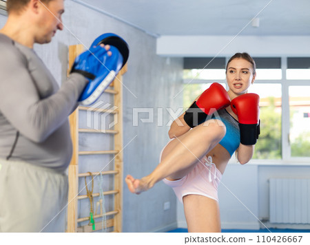 Athletic woman in boxing gloves kicks the punch mitts held by trainer Athletic woman in boxing gloves kicks the punch mitts held by trainer 110426667