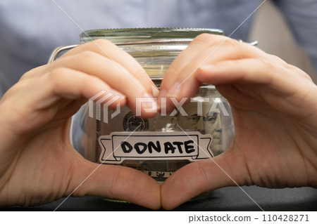 Female showing heart sign with hands Saving Money In Glass Jar filled with Dollars banknotes. DONATE transcription in front of jar. Managing personal finances extra income for future insecurity 110428271