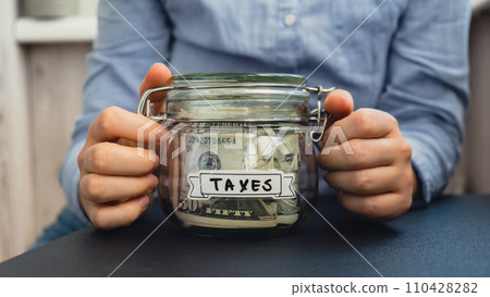 Female hands holding Glass jar full of American currency dollars cash banknote with text TAXES. Preparation saving money. Moderate consumption and economy Female hands holding Glass jar full of American currency dollars cash banknote with text TAXES. Preparation saving money. Moderate consumption and economy 110428282
