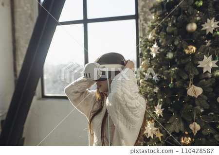 With sunlight streaming in, a vibrant young woman uses a virtual reality headset on a winter morning. With sunlight streaming in, a vibrant young woman uses a virtual reality headset on a winter morning. 110428587