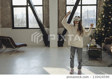 In the brightness of her apartment, a lively young woman wears a virtual reality headset. 110428590