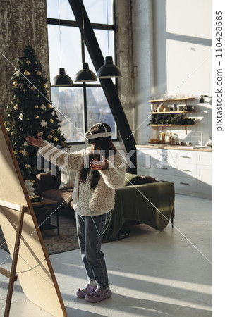 In the midst of Christmas cheer, a young lady in light attire and a VR headset admires herself in the mirror. In the midst of Christmas cheer, a young lady in light attire and a VR headset admires herself in the mirror. 110428685