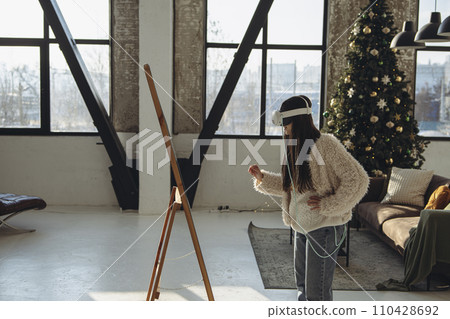 With the holiday ambiance prevailing, a young woman in light attire and a VR headset gazes into the mirror. With the holiday ambiance prevailing, a young woman in light attire and a VR headset gazes into the mirror. 110428692