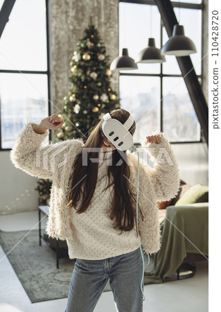 In front of a Christmas tree, a lovely young lady wears a virtual reality headset. 110428720