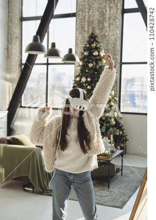 Embracing the festive spirit, a beautiful young lady uses a VR headset near a Christmas tree. Embracing the festive spirit, a beautiful young lady uses a VR headset near a Christmas tree. 110428742