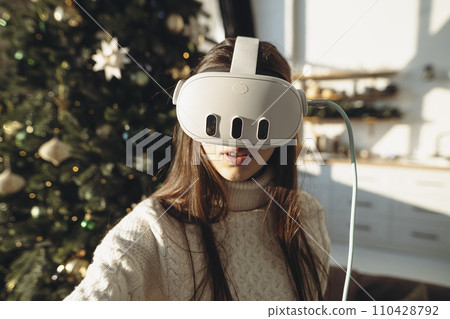 An image of a lovely girl wearing a virtual reality headset against the background of a Christmas tree. An image of a lovely girl wearing a virtual reality headset against the background of a Christmas tree. 110428792