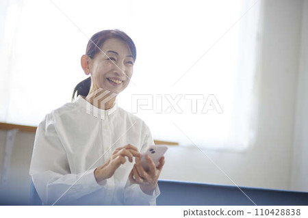 一位日本老婦人在客廳看著她的智慧型手機 一位日本老婦人在客廳看著她的智慧型手機 110428838