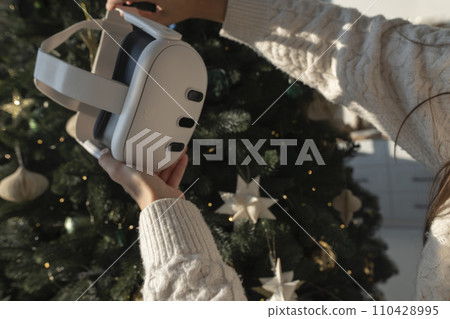 In front of a Christmas tree, a girl is holding a virtual reality headset. In front of a Christmas tree, a girl is holding a virtual reality headset. 110428995