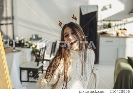 In the brightness of her apartment, a lively young woman wears a Christmas reindeer mask. In the brightness of her apartment, a lively young woman wears a Christmas reindeer mask. 110428996