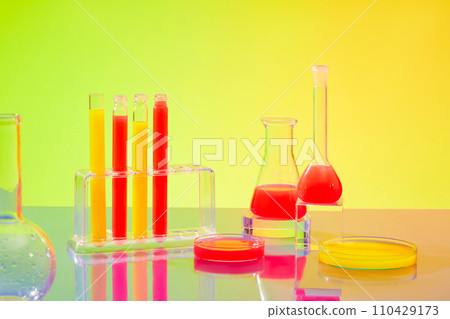 Lively hues fill test tubes, petri dishes, beakers, and Erlenmeyer flasks set against a green-yellow gradient backdrop within the laboratory environment. 110429173