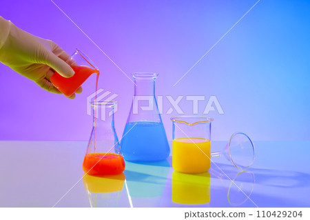 A hand is holding a beaker pouring red liquid from it through an erlenmeyer flask, next to it are two experimental glass flasks containing yellow and blue liquids on a gradient background. 110429204