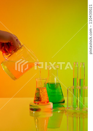 Hand model holding a cup pouring orange liquid into a small erlenmeyer flask. A rack of test tubes featured. Science laboratory research and development concept 110430021