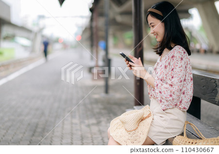 young asian woman traveler with weaving basket using a mobile phone and waiting for train in train station. Journey trip lifestyle, world travel explorer or Asia summer tourism concept. 110430667