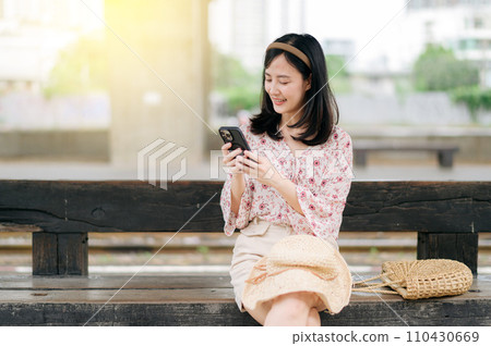 young asian woman traveler with weaving basket using a mobile phone and waiting for train in train station. Journey trip lifestyle, world travel explorer or Asia summer tourism concept. 110430669