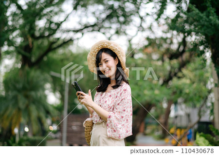 Portrait of asian young woman traveler with weaving hat and basket using mobile phone on green park nature background. Journey trip lifestyle, world travel explorer or Asia summer tourism concept. Portrait of asian young woman traveler with weaving hat and basket using mobile phone on green park nature background. Journey trip lifestyle, world travel explorer or Asia summer tourism concept. 110430678