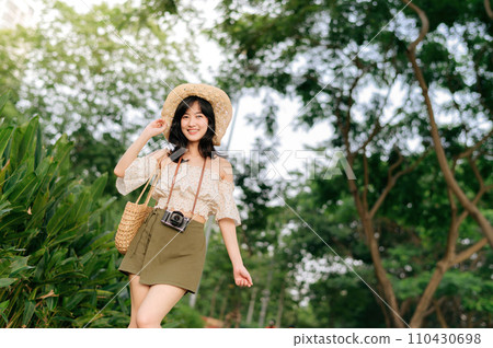 Portrait of asian young woman traveler with weaving hat and basket and a camera on green public park nature background. Journey trip lifestyle, world travel explorer or Asia summer tourism concept. Portrait of asian young woman traveler with weaving hat and basket and a camera on green public park nature background. Journey trip lifestyle, world travel explorer or Asia summer tourism concept. 110430698