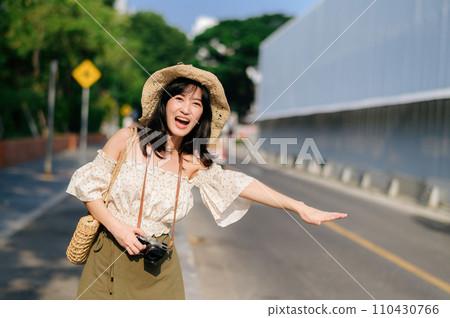 Portrait of asian young woman traveler with weaving hat and basket and a camera waving hand to friend by the street. Journey trip lifestyle, world travel explorer or Asia summer tourism concept. Portrait of asian young woman traveler with weaving hat and basket and a camera waving hand to friend by the street. Journey trip lifestyle, world travel explorer or Asia summer tourism concept. 110430766