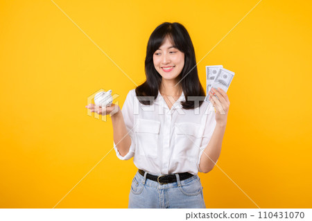 Portrait beautiful young business asian woman wearing white shirt and denim jean with a lot of cash money and piggy bank isolated on yellow background. Wealth money saving, finance investment concept. 110431070
