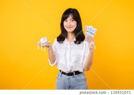 Portrait beautiful young business asian woman wearing white shirt and denim jean with a lot of cash money and piggy bank isolated on yellow background. Wealth money saving, finance investment concept. 110431071