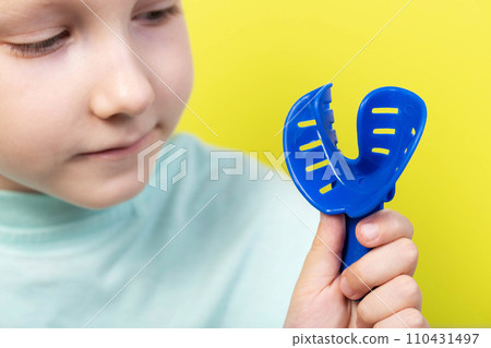 A seven year old girl looks at a blue dental impression tray in orthodontics. Concept for manufacturing braces and dentures for children, correction of malocclusion. Yellow background, dentistry 110431497