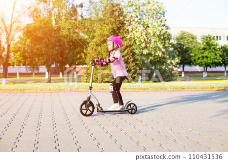 A beautiful girl of 7 years old rides a scooter in a pink protection against injuries and falls. Sunny summer day. Copy space for text A beautiful girl of 7 years old rides a scooter in a pink protection against injuries and falls. Sunny summer day. Copy space for text 110431536