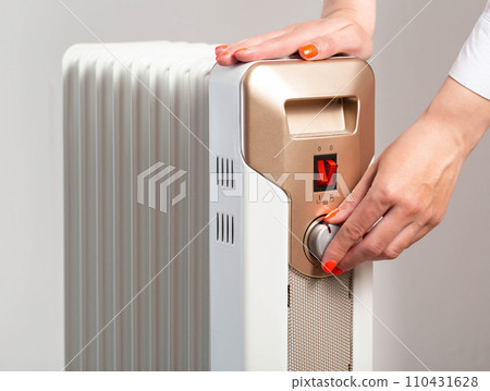 A woman s hand with a beautiful manicure regulates the room heating temperature on an electric heater. The concept of turning on the heating in the autumn-winter period. Cold in apartments, close-up A woman s hand with a beautiful manicure regulates the room heating temperature on an electric heater. The concept of turning on the heating in the autumn-winter period. Cold in apartments, close-up 110431628