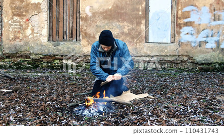 Homeless man sits on ground and pushes pieces of cardboard into fire to keep warm from cold. Tramp in dirty clothes and hat cap. Below poverty line. Illegal immigrant. Male refugee. Fugitive beggar 110431743